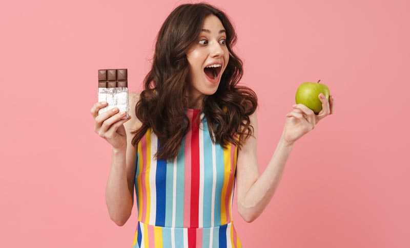 Überraschte Frau in gestreiftem Kleid hält Schokolade und Apfel in den Händen und schaut auf den Apfel. Symbol für gesunde Entscheidung und Zuckerverzicht.
(c) AdobeStock