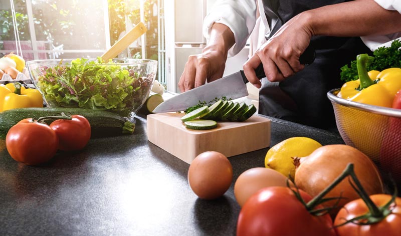Adipositas-Therapie im Realitätscheck: Warum GLP-1 alleine uns nicht rettet 1 Nahaufnahme in einer Küche, wo ein Koch mit einem großen Messer Zucchini auf einem Holzbrett schneidet, umgeben von frischen Tomaten, Paprika, Zwiebeln und Salat, Stichwort Adipositas-Therapie.
(c) AdobeStock