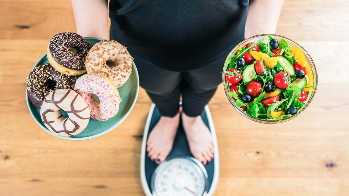 Adipositas-Therapie im Realitätscheck: Warum GLP-1 alleine uns nicht rettet Blick von oben auf eine Frau auf einer Personenwaage, die in der einen Hand einen Teller mit süßen Donuts und in der anderen eine gesunde Schüssel Salat hält. (c) AdobeStock