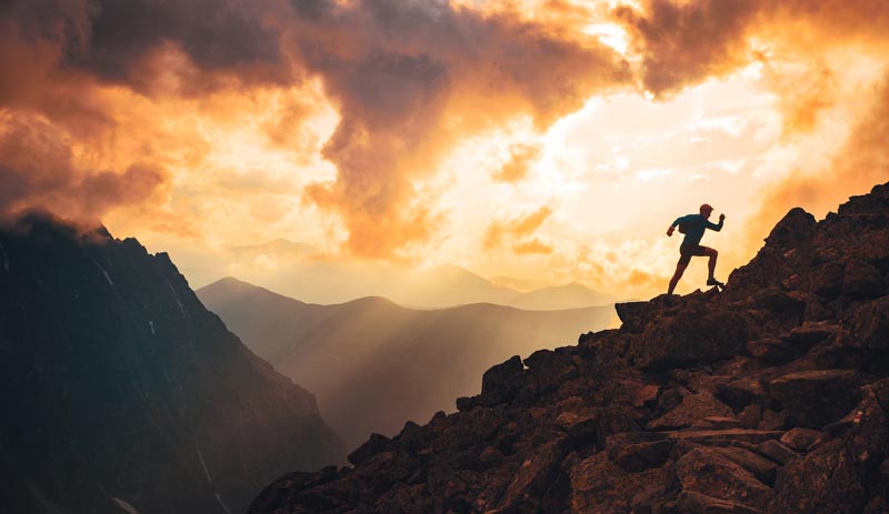 Körperliche Souveränität: Warum Fitness nicht aus der Apotheke kommt 2 Silhouette eines Läufers, der im spektakulären, goldenen Sonnenuntergang einen extrem steilen, felsigen Berggipfel erklimmt. Im Hintergrund sind Bergketten und dramatische Wolken zu sehen, Stichwort Körperliche Souveränität.
(c) AdobeStock
