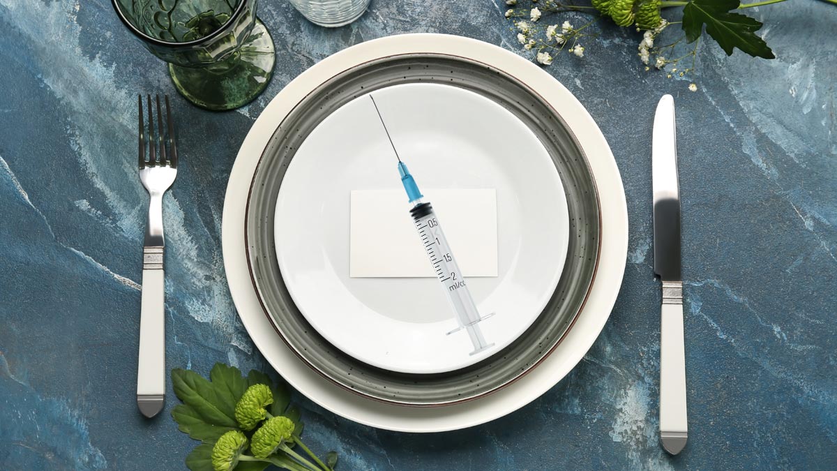 Körperliche Souveränität: Warum Fitness nicht aus der Apotheke kommt Ein elegant gedeckter blauer Tisch mit weißem Porzellanteller, Silberbesteck, Gläsern und grünen Blumen. Auf dem Teller liegt eine sterile, leere Spritze auf einer weißen Karte. (c) AdobeStock/ Alterneudenken