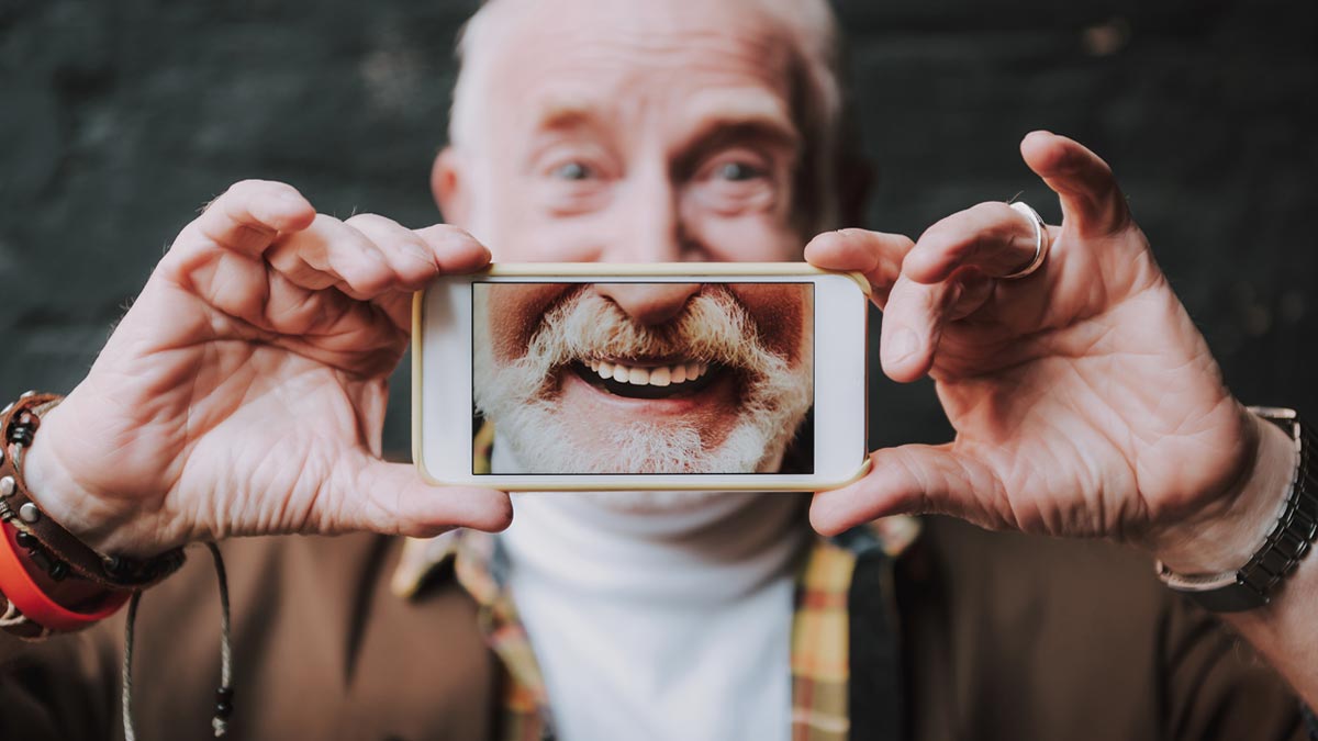 Cyberangriffe auf ältere Menschen: Die neue Dimension der digitalen Jagd Älterer Mann hält sich ein Smartphone vor das Gesicht, auf dessen Bildschirm ein lachender Mund mit weißem Bart zu sehen ist, Symbol für digitale Identitätsfälschung und Deepfakes. (c) AdobeStock