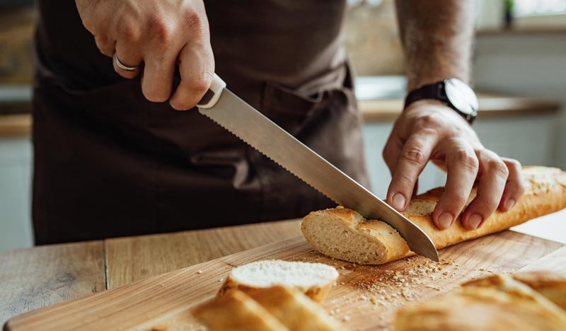 Nahaufnahme von Männerhänden in einer braunen Schürze, die mit einem gezackten Brotmesser ein Baguette auf einem Holzschneidebrett in Scheiben schneiden.
(c) AdobeStock