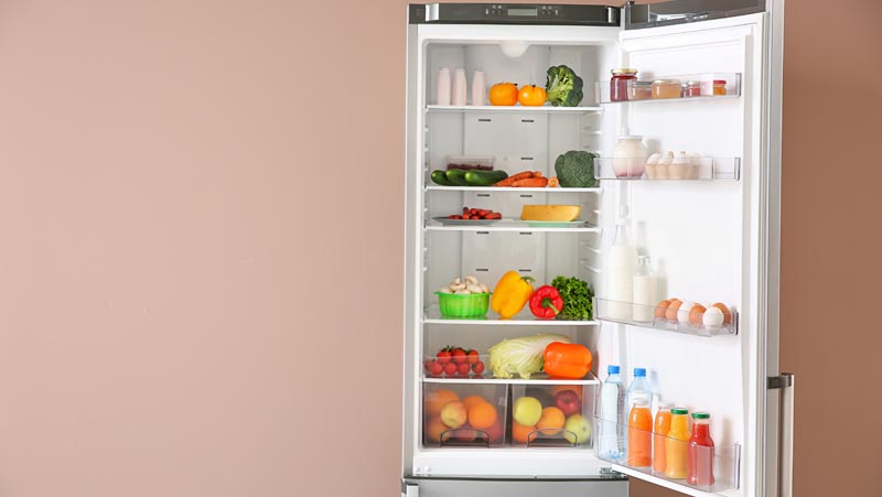 Ein komplett geöffneter, sauber strukturierter und heller Kühlschrank, der reichlich mit frischem Gemüse, Obst und abgefüllten Flaschen vor einer beigen Wand gefüllt ist, Stichwort Lebensmittelverschwendung.
(c) AdobeStock