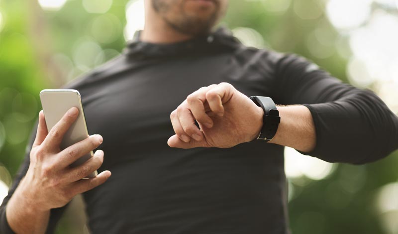 Nahaufnahme eines Mannes in Sportkleidung, der gleichzeitig auf sein Smartphone und seine Smartwatch am Handgelenk blickt.
(c) AdobeStock