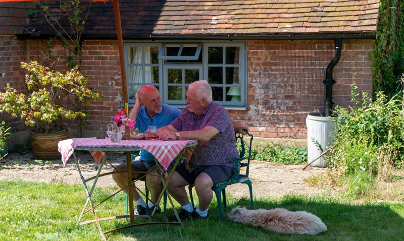 Zwei ältere Männer sitzen entspannt an einem einfachen Gartentisch vor einem Backsteinhaus, ein Hund liegt im Gras daneben; Stichwort Lüge vom aktiven Altern.
(c) AdobeStock