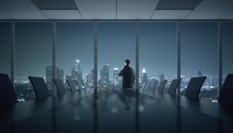 Silhouette eines Mannes im Anzug von hinten, der nachts in einem leeren, dunklen Sitzungssaal am Telefon telefoniert und auf eine hell erleuchtete Skyline blickt.
(c) AdobeStock
