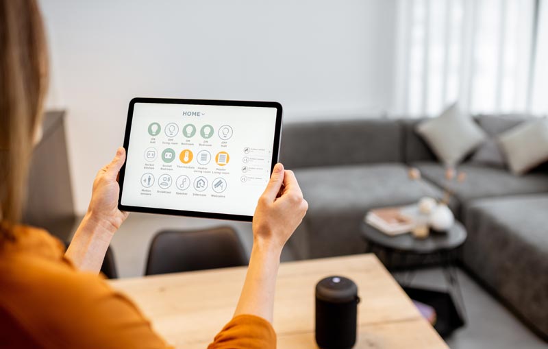 Person hält ein Tablet mit einer Smart-Home-Steuerungs-App in einem modern eingerichteten Wohnzimmer, Stichwort Altersdiskriminierung.
(c) AdobeStock