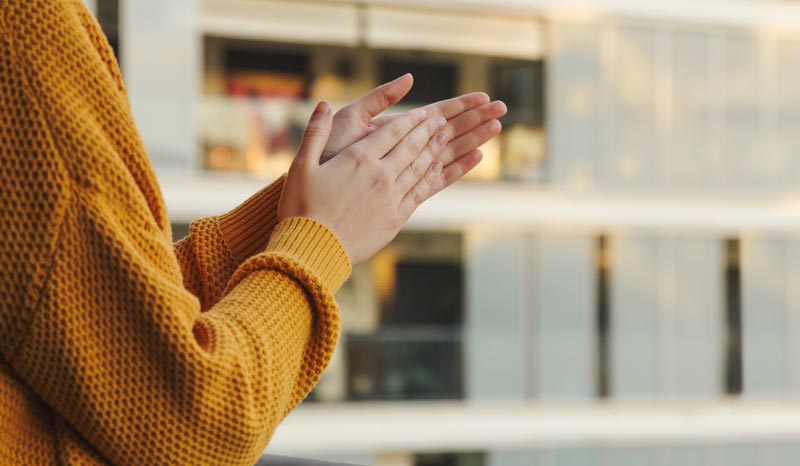 Eine Person in einem gelben Strickpullover steht auf einem Balkon vor einem modernen Wohngebäude und klatscht in die Hände, Stichwort Tag der Pflege Kritik.
(c) AdobeStock