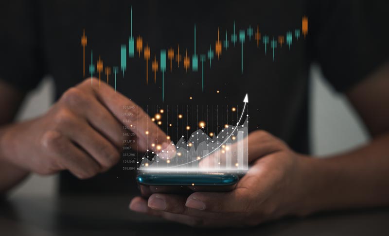 Eine Hand hält ein Smartphone, über dem leuchtende Finanzcharts und eine Wachstumskurve als Symbol für Profit durch den Alters-Algorithmus beim Online-Shopping schweben.(c) AdobeStock