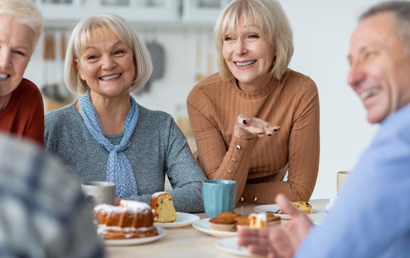 Eine gut gelaunte Seniorengruppe beim Kaffeeklatsch. Sie sitzen lachend um einen Tisch voller Kuchen und Muffins, StichwortAlters-Einsamkeit Digital vernetzt.(c) AdobeStock