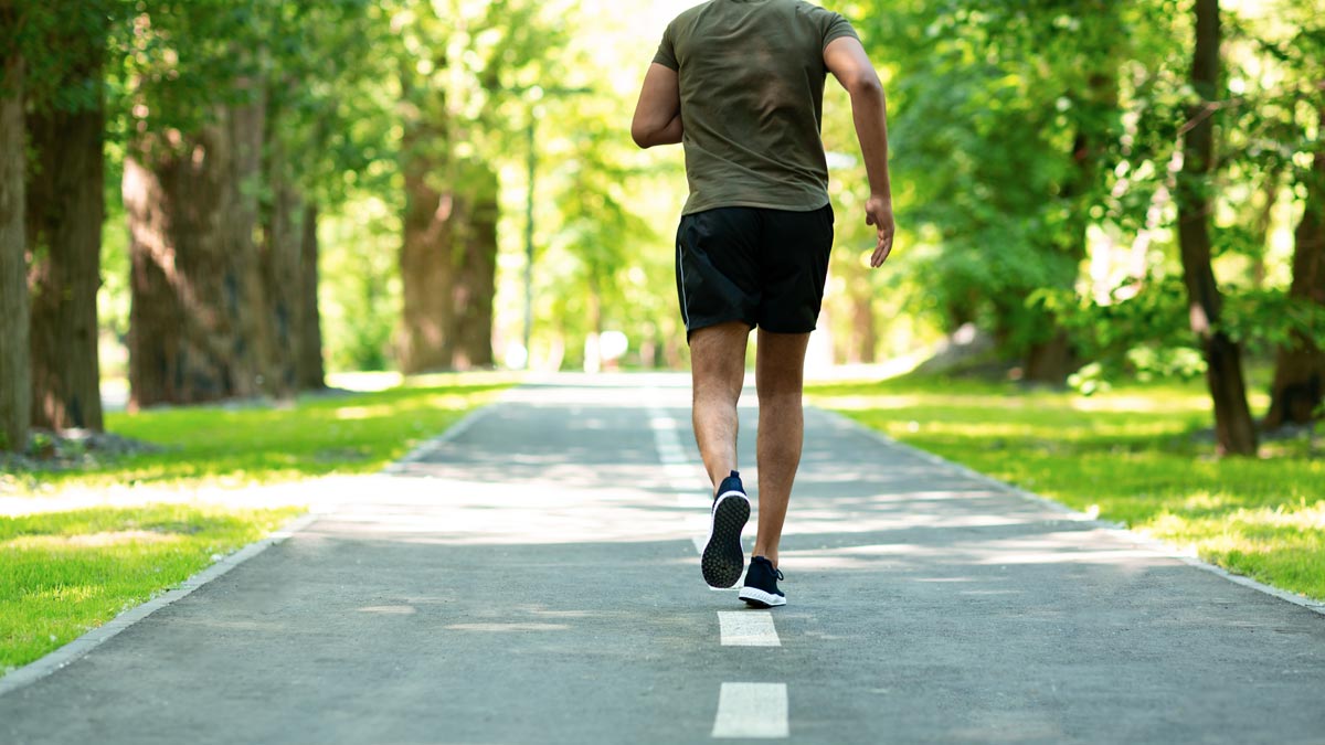 Barfußschuhe als orthopädisches Risiko? Segen und Fluch des Trends Ein Mann von hinten beim Joggen auf einem asphaltierten Parkweg, der von dichten, grünen Bäumen gesäumt ist. (c) AdobeStock