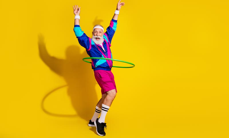 Ein älterer, bärtiger Mann im bunten 80er-Jahre-Trainingsanzug kreist gut gelaunt einen grünen Hula-Hoop-Reifen vor gelbem Hintergrund, Stichwort High Protein Trend.(c) AdobeStock