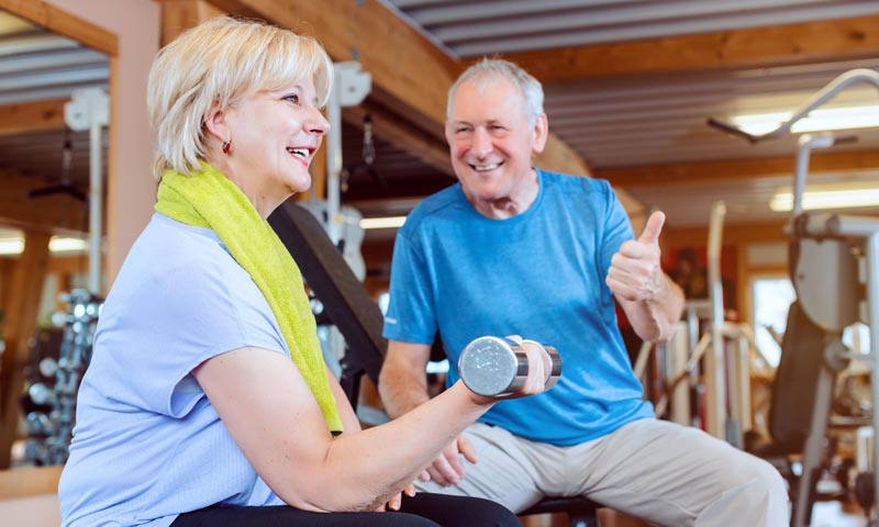 Eine ältere Frau mit blonden Haaren trainiert im Fitnessstudio mit einer Hantel, während im Hintergrund ein älterer Mann auf einer Bank sitzt und lächelnd den Daumen nach oben zeigt, Stichwort Insulinresistenz.(c) AdobeStock