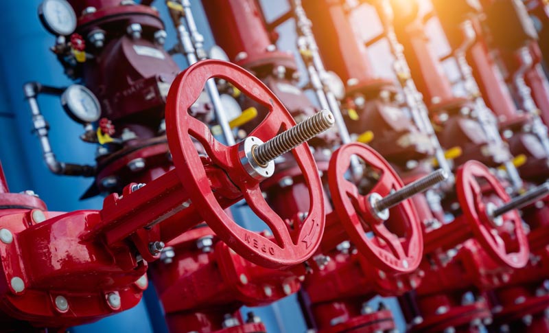 Eine Reihe großer, roter Absperrventile und Drehräder an industriellen Rohrleitungen vor blauem Hintergrund im Profil.(c) AdobeStock