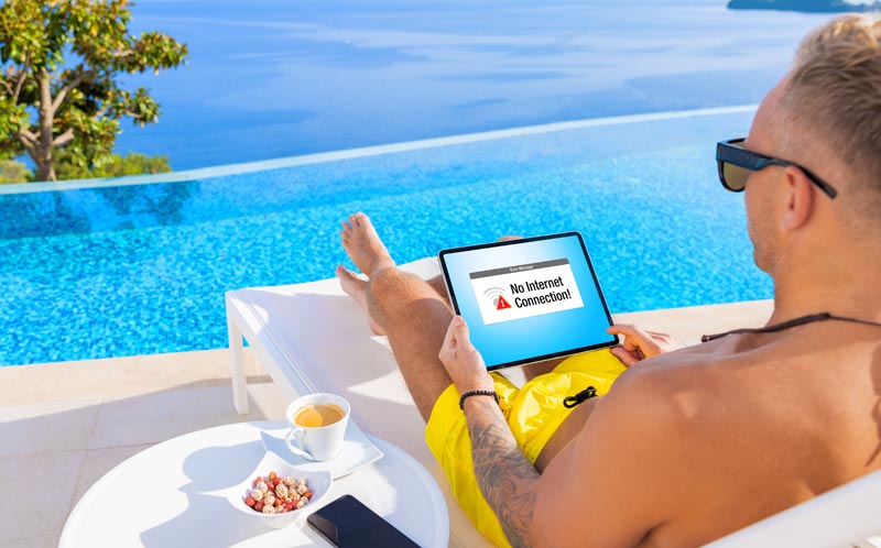 Roaming-Fallen im Urlaub: Die schmerzhafte Wahrheit über Smartphone-Mythen 2 Ein Mann liegt auf einer Sonnenliege am Infinity-Pool mit Blick aufs Meer und hält ein Tablet, das die Fehlermeldung „No Internet Connection!“ anzeigt.(c) AdobeStock