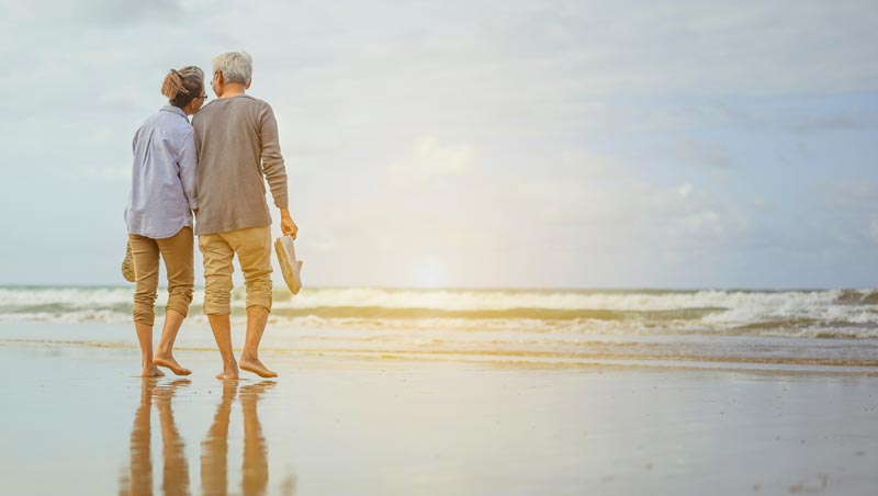 Ein älteres Paar läuft barfuß und Hand in Hand an einem sonnigen Sandstrand entlang ins flache Meerwasser.(c) AdobeStock