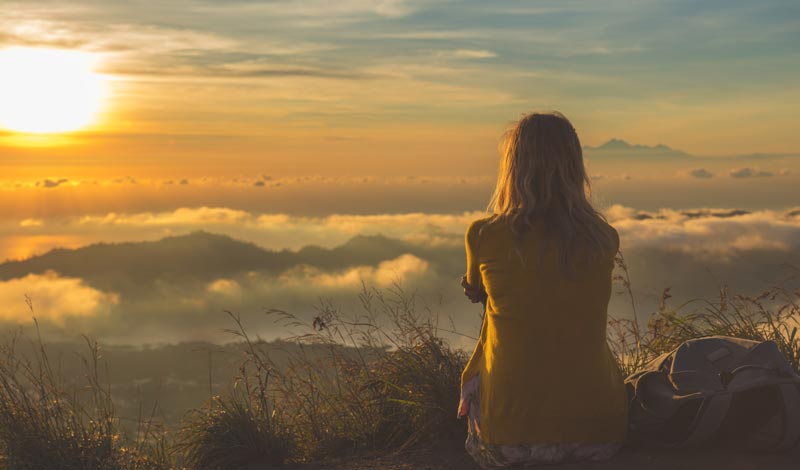 Eine Frau von hinten mit blonden Haaren und gelber Jacke sitzt auf einem Berggipfel und blickt auf ein Wolkenmeer während des Sonnenuntergangs.
(c) AdobeStock