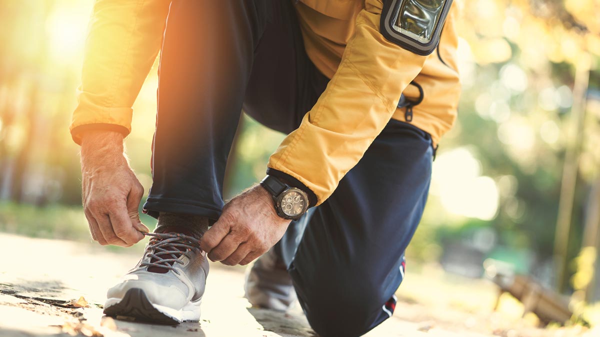 Wearables – Alter, Herzwerte und Fitness im Ü50-Check Nahaufnahme eines älteren Läufers in gelber Sportjacke, der sich im Freien hinkniet, um seine Laufschuhe zu binden, mit einer Sportuhr am Handgelenk. (c) AdobeStock