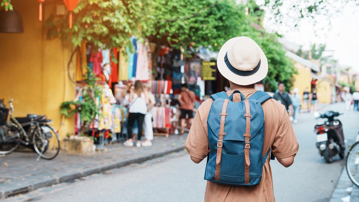 Radikale Selbsterkenntnis: Warum Sie alleine reisen im Alter als Ego-Stresstest nutzen sollten Ein Reisender mit Hut und blauem Rucksack steht in einer fremden asiatischen Straße und nutzt das alleine reisen im Alter als kognitiven Stresstest. (c) AdobeStock