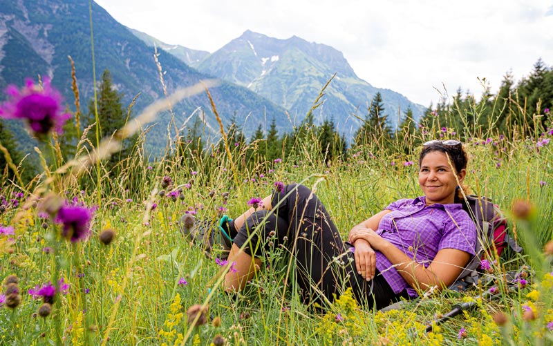 Eine entspannte Wanderin liegt lächelnd in einer blühenden Almwiese vor einer Bergkulisse in Österreich.
(c) AdobeStock
