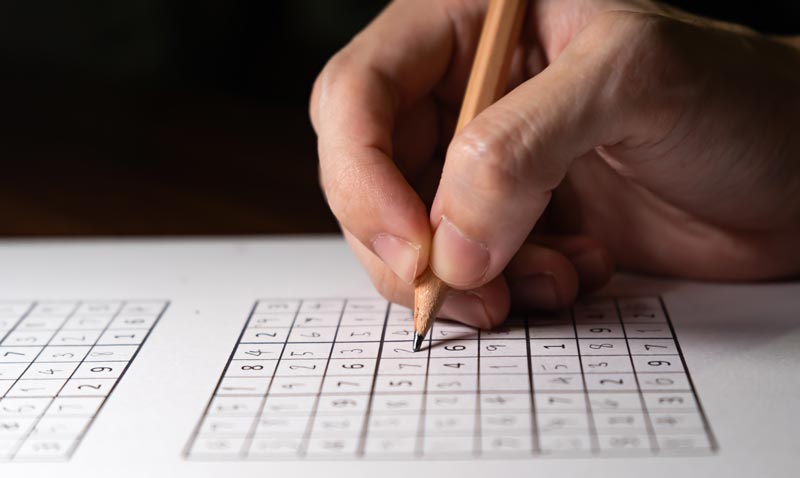 Gehirn fit halten im Alter: Smartphone schlägt Sudoku 1 Nahaufnahme einer Hand, die mit einem Bleistift ein Sudoku-Rätsel ausfüllt, statt das Gehirn fit halten im Alter durch komplexe digitale Reize zu fördern.(c) AdobeStock