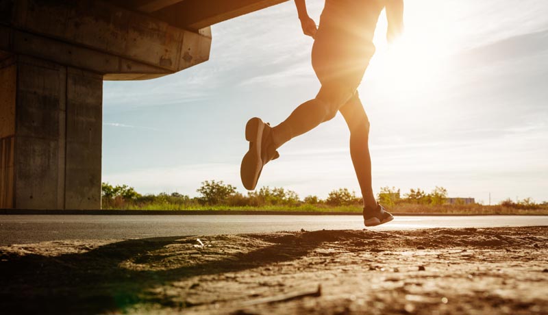 Dynamische Nahaufnahme der Beine eines Läufers im hellen Sonnenlicht auf dem Asphalt als Symbol für Bewegung und gesund bleiben im Alter.(c) AdobeStock