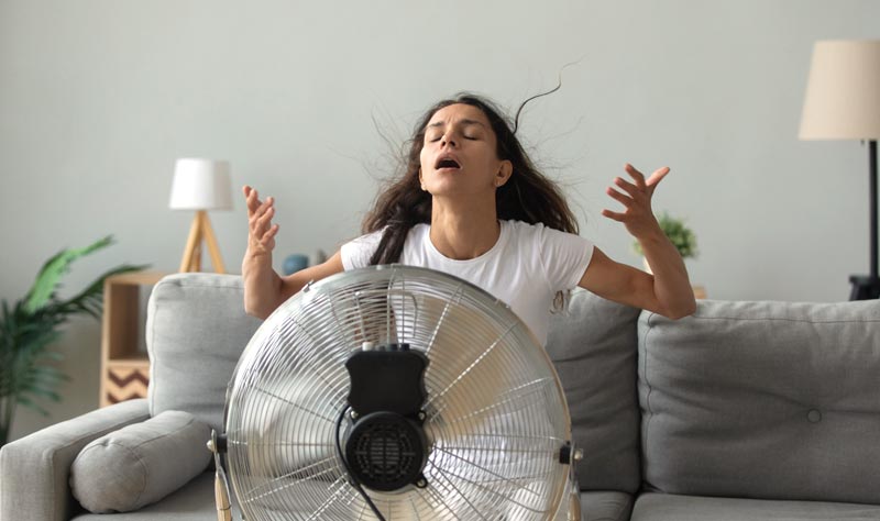 Eine Frau sitzt mit dramatisch geöffnetem Mund und fliegenden Haaren direkt vor einem großen, laufenden Standventilator auf dem Sofa.(c) AdobeStock