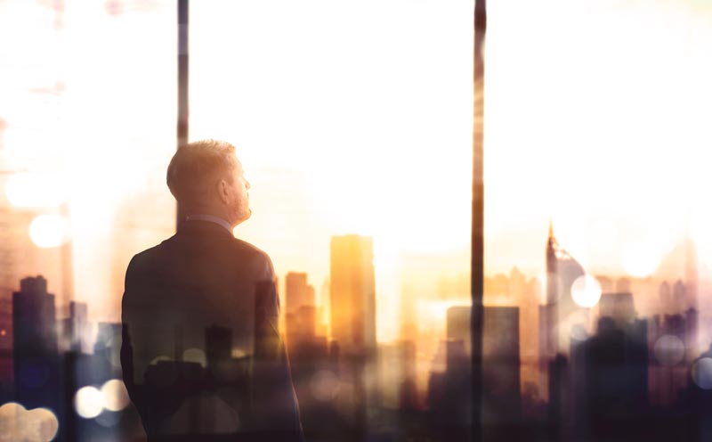 Silhouette eines Mannes im Anzug von hinten, der aus einem großen Fenster im Hochhaus auf eine Großstadt-Skyline im hellen Sonnenuntergang blickt.(c) AdobeStock