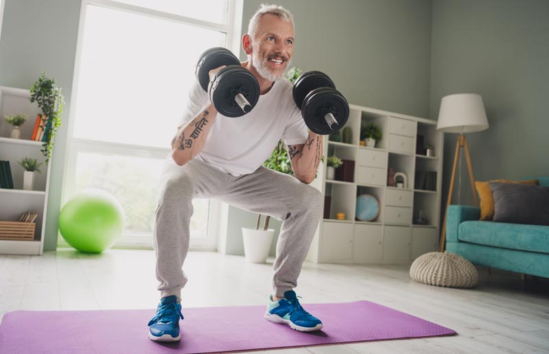 Ein fitter, grauhaariger Mann im reifen Alter absolviert lächelnd Kniebeugen mit zwei schweren Kurzhanteln auf einer lila Trainingsmatte zu Hause, Stichwort Männergesundheit ab 50.(c) AdobeStock