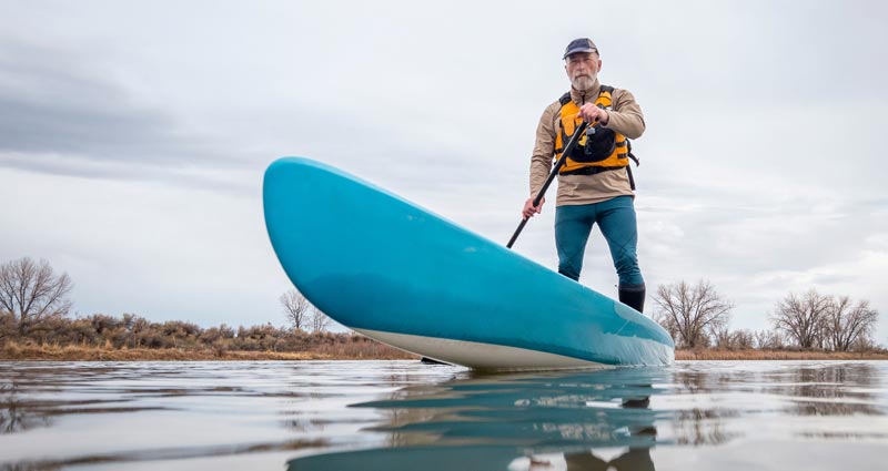 Ein älterer Mann mit grauem Bart steht im Winteroutfit mit Schwimmweste auf einem hellblauen Stand-Up-Paddle-Board auf einem ruhigen Fluss unter bewölktem Himmel.
(c) AdobeStock