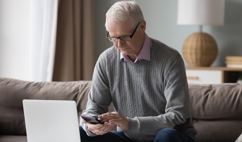 Ein älterer Mann mit Brille und grauem Pullover sitzt auf einem Sofa vor einem Laptop und bedient konzentriert ein Smartphone, Stichwort Marketing-Blase.
(c) AdobeStock