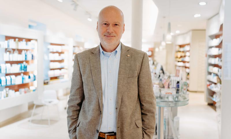 Mag. Martin R. Geisler, Generalsekretär der ApoLife Apothekengruppe, lächelt im Sakko in einer hell erleuchteten, modernen Apotheke.(c) ApoLife