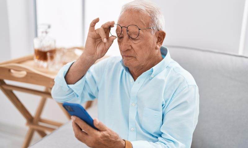 Ein älterer Mann sitzt auf einer Couch, hebt seine Brille an und blickt skeptisch auf sein Handy, um sich vor der Quishing Betrugsmasche zu schützen.(c) AdobeStock