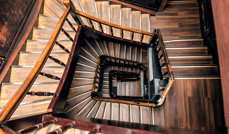 Blick von oben in das tiefe, eckige Auge einer alten Holztreppe mit kunstvollem Geländer.(c) AdobeStock