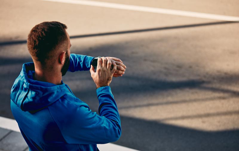 Rückansicht eines Mannes in blauer Sportjacke, der auf seine Smartwatch am Handgelenk blickt, um Trainingsdaten wie Herzfrequenz oder VO2max zu überprüfen, Stichwort Vo2max im Alter.(c) AdobeStock