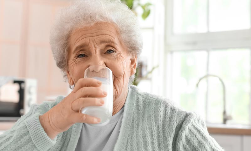 Longevity-Nährstoffe: Die ewige Jugend aus dem Becher oder reine Geldmacherei? 1 Eine lächelnde, ältere Frau mit grauem Haar trinkt in einer hellen Küche ein Glas weiße Flüssigkeit (Milch oder Kefir).(c) AdobeStock
