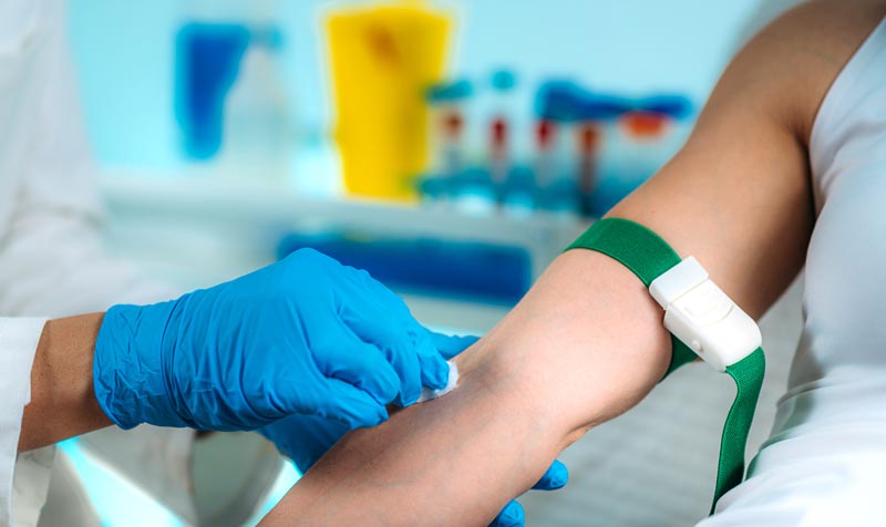 Eine medizinische Fachkraft mit blauen Handschuhen desinfiziert den Arm einer Patientin vor einer Blutentnahme.(c) AdobeStock