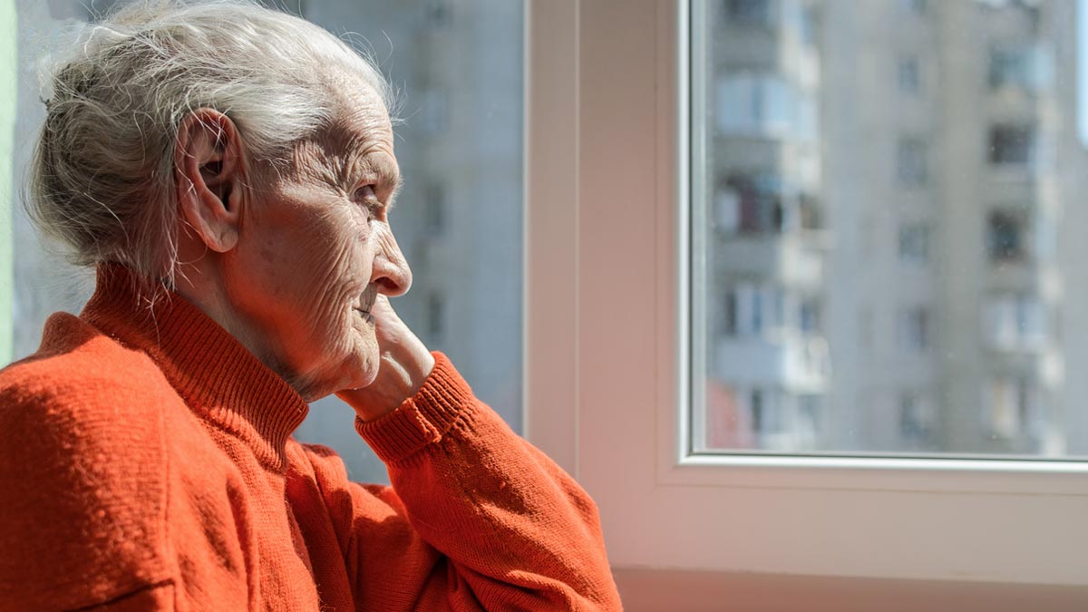 „Soziale Sarkopenie“: Warum Einsamkeit im Alter so gefährlich ist wie 15 Zigaretten am Tag Ältere Frau mit rotem Pullover blickt stumm und nachdenklich aus dem Fenster einer Hochhauswohnung. (c) AdobeStock