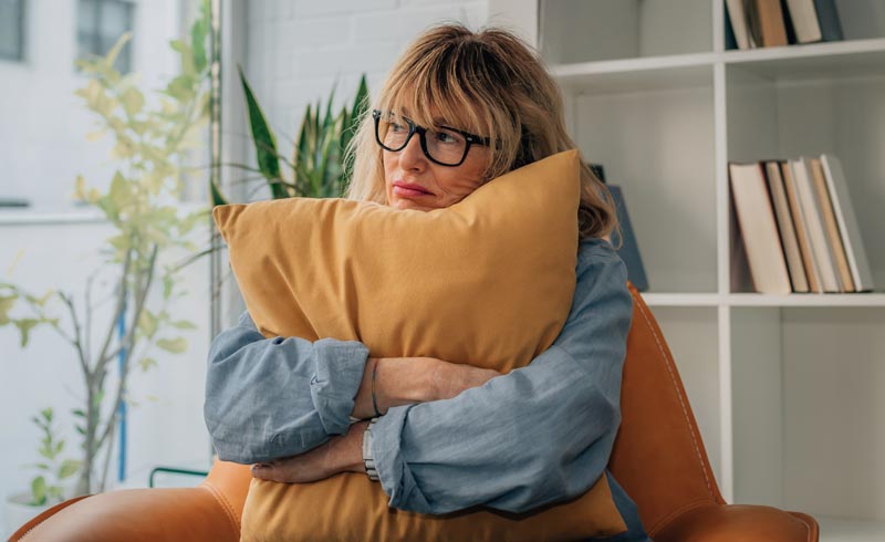 Eine reife Frau mit Brille sitzt nachdenklich im Sessel und umarmt ein gelbes Kissen, Stichwort Darmgesundheit im Alter.(c) AdobeStock