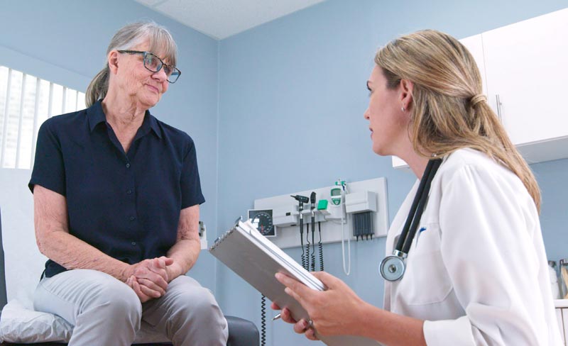 Eine ältere Patientin im Arztzimmer im Gespräch mit einer Ärztin, die eine Klemmmappe hält.(c) AdobeStock