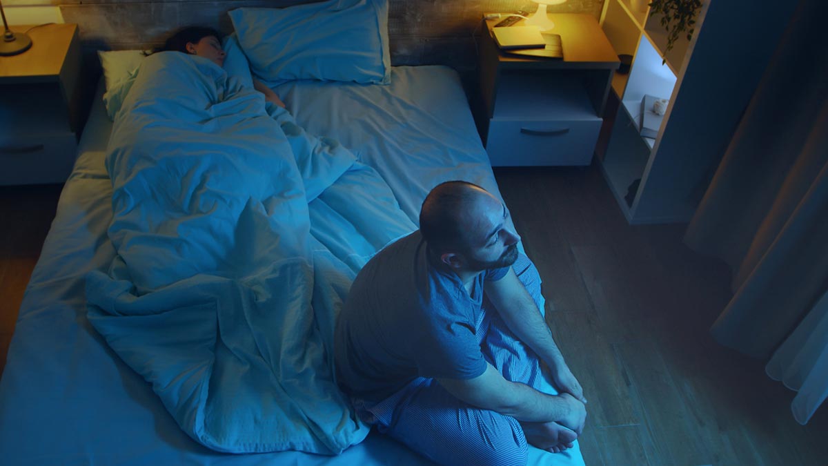 Schlafstörungen ab 50: Warum 8 Stunden Schlaf ein Mythos sind (und was wirklich hilft) Mann sitzt nachts mit Schlafstörungen auf der Bettkante, während im Hintergrund seine Partnerin schläft. (c) AdobeStock