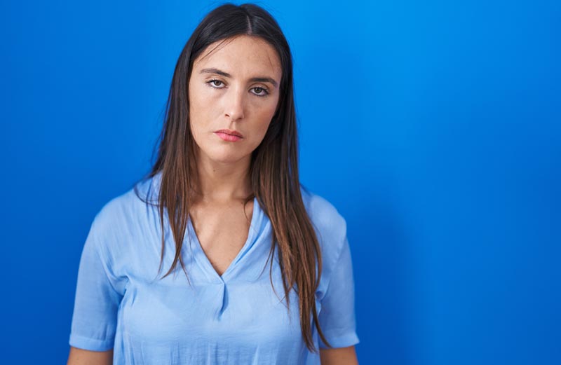 Frau blickt erschöpft und frustriert direkt in die Kamera vor blauem Hintergrund.(c) AdobeStock