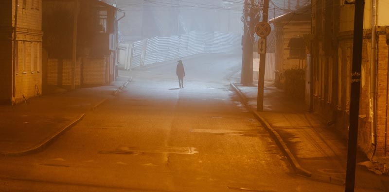 Einsam gehende Person aus der Ferne in einer düsteren, orange beleuchteten und nebligen Straße bei Nacht.(c) AdobeStock