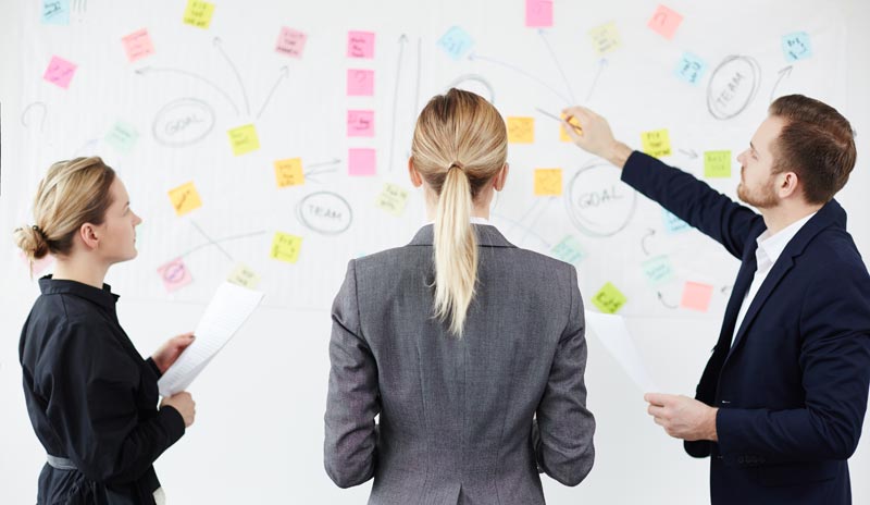 Dreiköpfiges Büro-Team steht vor einem Board voller bunter Post-its und Zettel und diskutiert Prozesse, Stichwort Pragmatismus im Beruf.(c) AdobbeStock