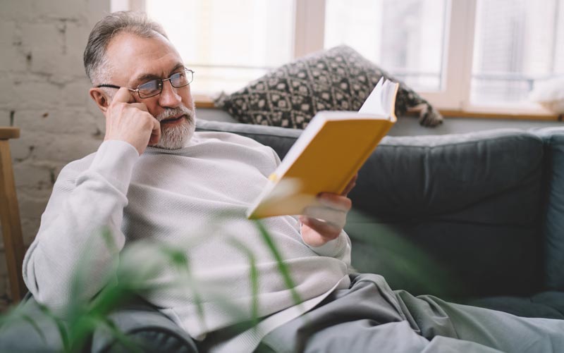 Älterer Mann mit Brille u. Graubart liegt entspannt auf einem Sofa und liest aufmerksam in einem Buch, Stichwort Longevity Wahn.(c) AdobeStock