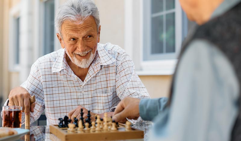 Zwei ältere Männer sitzen im Garten an einem Glasstisch u. spielen eine Partie Schach.(c) AdobeStock