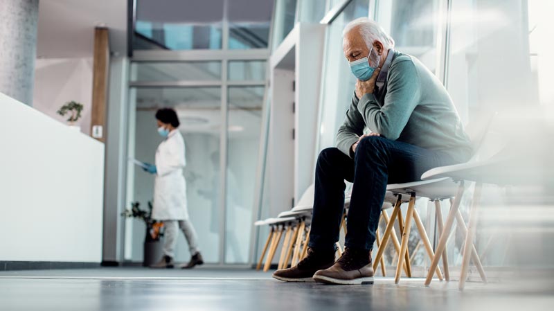 Migräne im Alter: Wenn moderne High-Tech-Medizin an der Kassenrealität scheitert 1 Ein älterer Mann mit Schutzmaske sitzt erschöpft und wartend auf einem Stuhl im Warteraum einer Klinik, Stichwort Migräne Behandlung.(c) AdobeStock