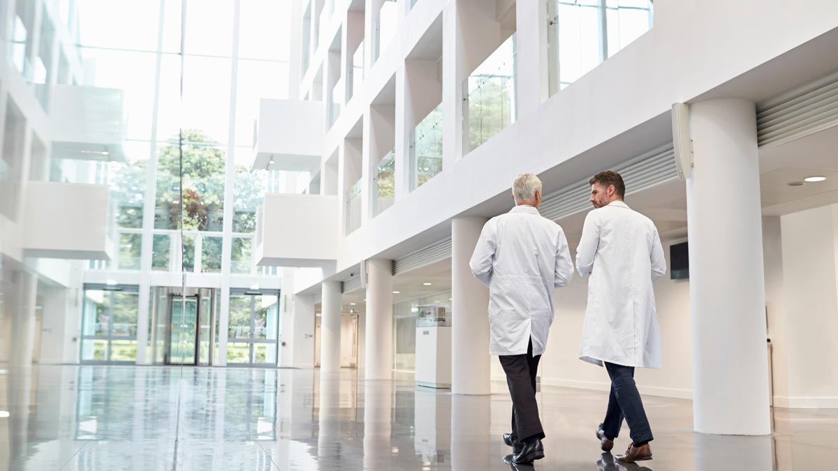 Vorsorgeuntersuchung Österreich: Das überfällige Evidenz-Update für Ihre Gesundheit Zwei Ärzte in weißen Mänteln gehen durch das lichtdurchflutete Atrium eines modernen Klinikgebäudes. (c) AdobeStock