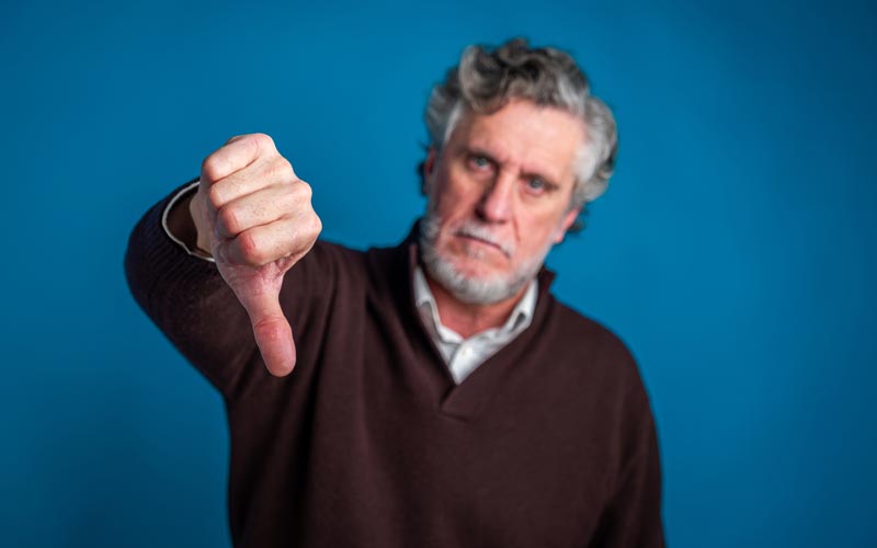 Älterer Mann mit grauem Haar und Bart streckt den Daumen nach unten vor blauem Hintergrund.   (c) AdobeStock